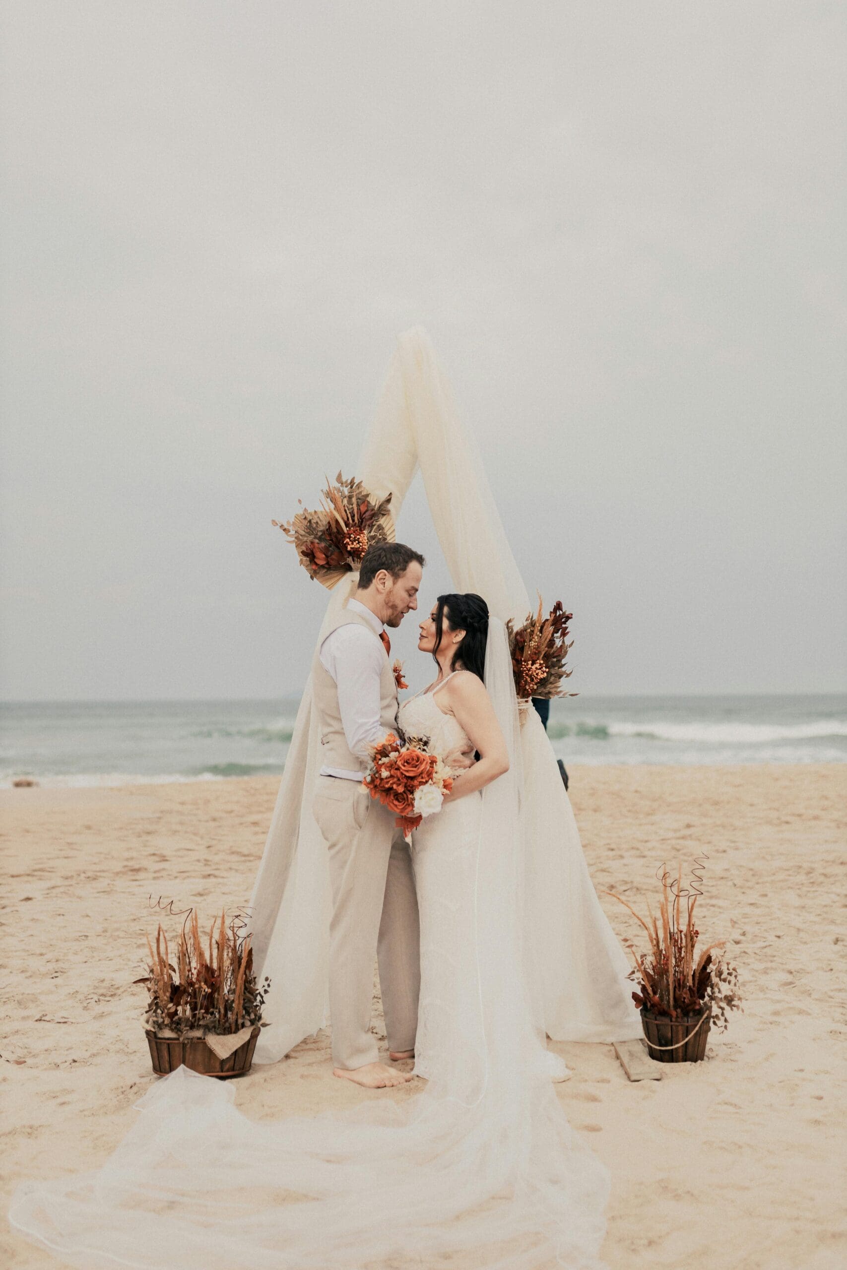 An elegant couple renews their vows in an intimate beach ceremony under a stylish bohemian arch, symbolizing a bespoke anniversary celebration.
