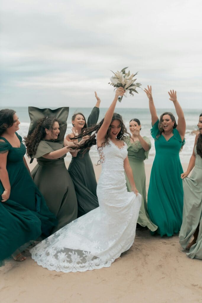 A group of happy bridesmaids in various shades of green dresses celebrate on a beach as the bride, holding her bouquet high, lifts her dress and smiles.