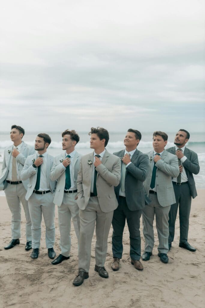 A group of six groomsmen in light gray and dark gray suits standing on a beach, adjusting their ties and posing for a photo.