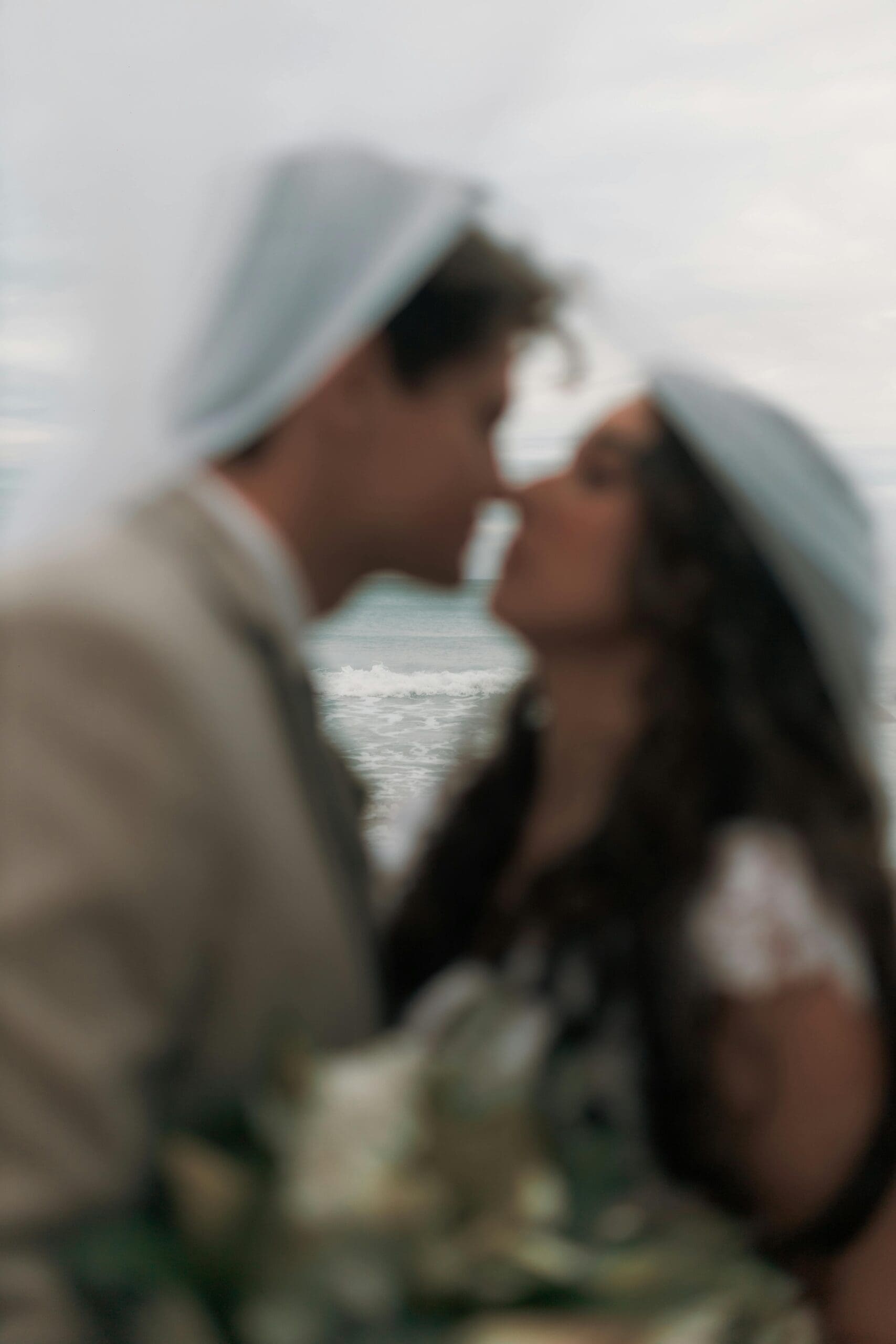 A close-up, out-of-focus photo of a couple kissing on a beach. The groom is wearing a beige jacket, and the bride has long, dark hair and a white lace dress. The ocean and sky are blurred in the background.