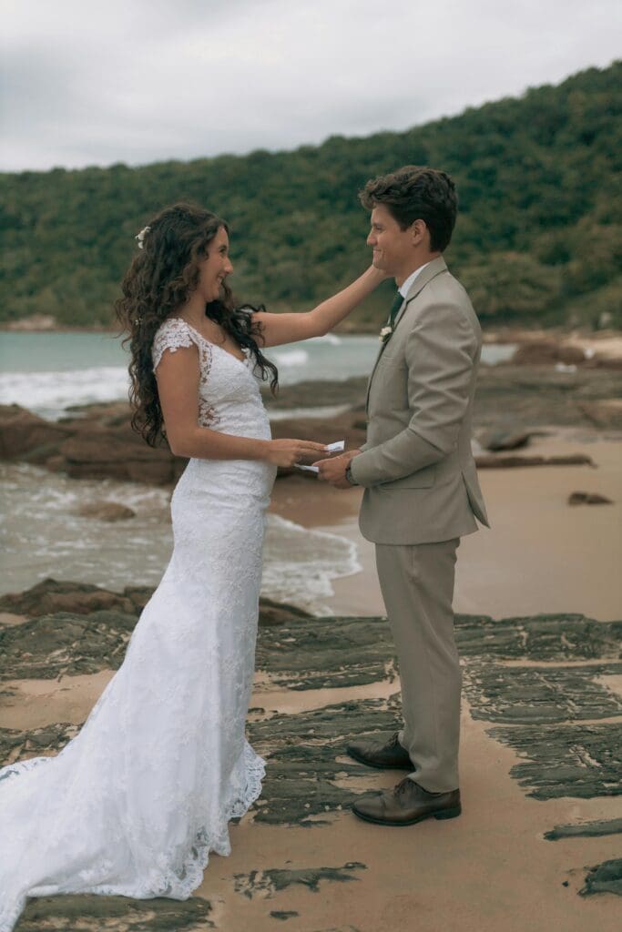 A newly married couple smiles at each other on a rocky beach. The bride, in a white lace dress, gently touches the groom's cheek as they hold hands.