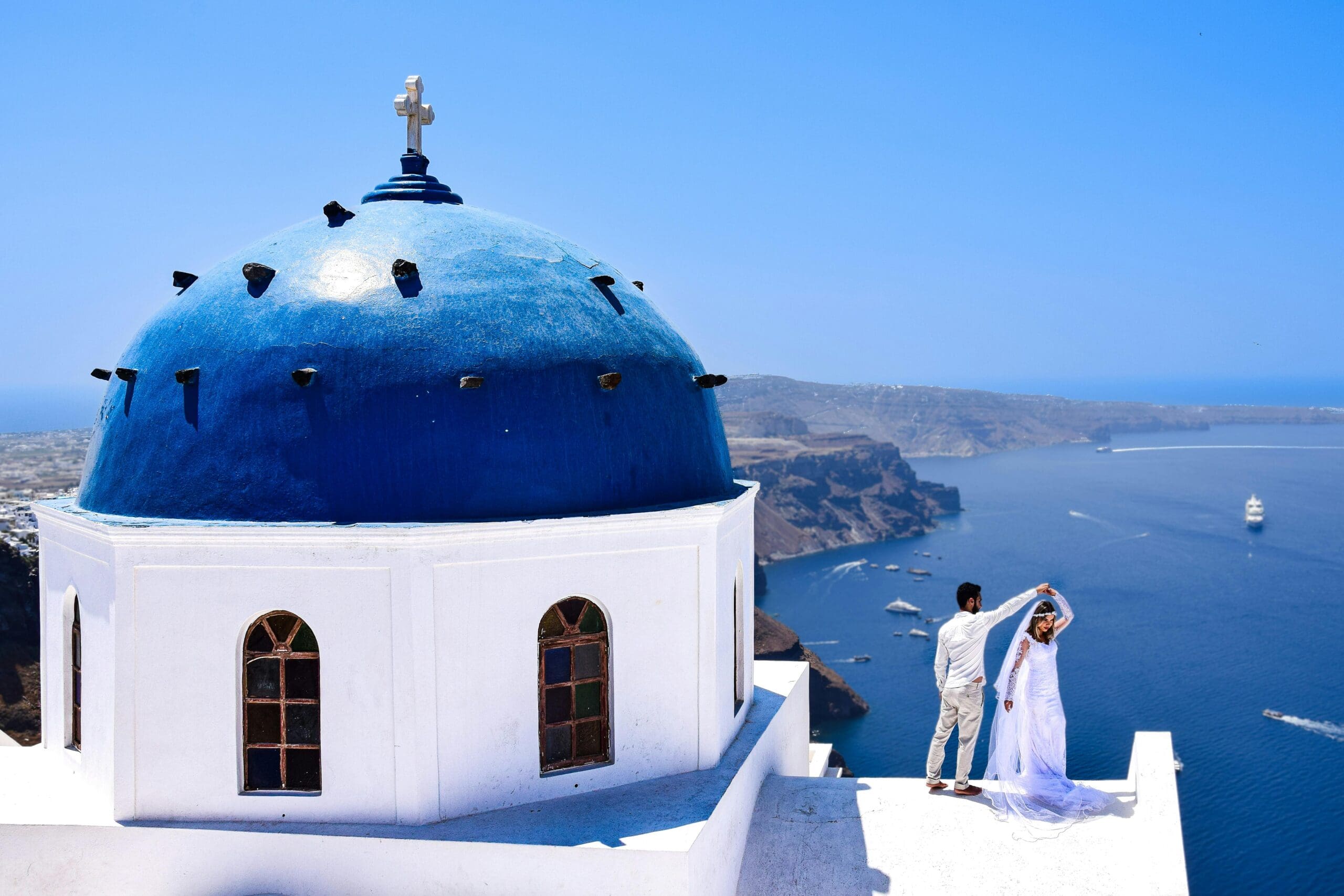 A newlywed couple on their honeymoon in Santorini, Greece, poses on a white rooftop next to an iconic blue-domed church overlooking the Aegean Sea.
