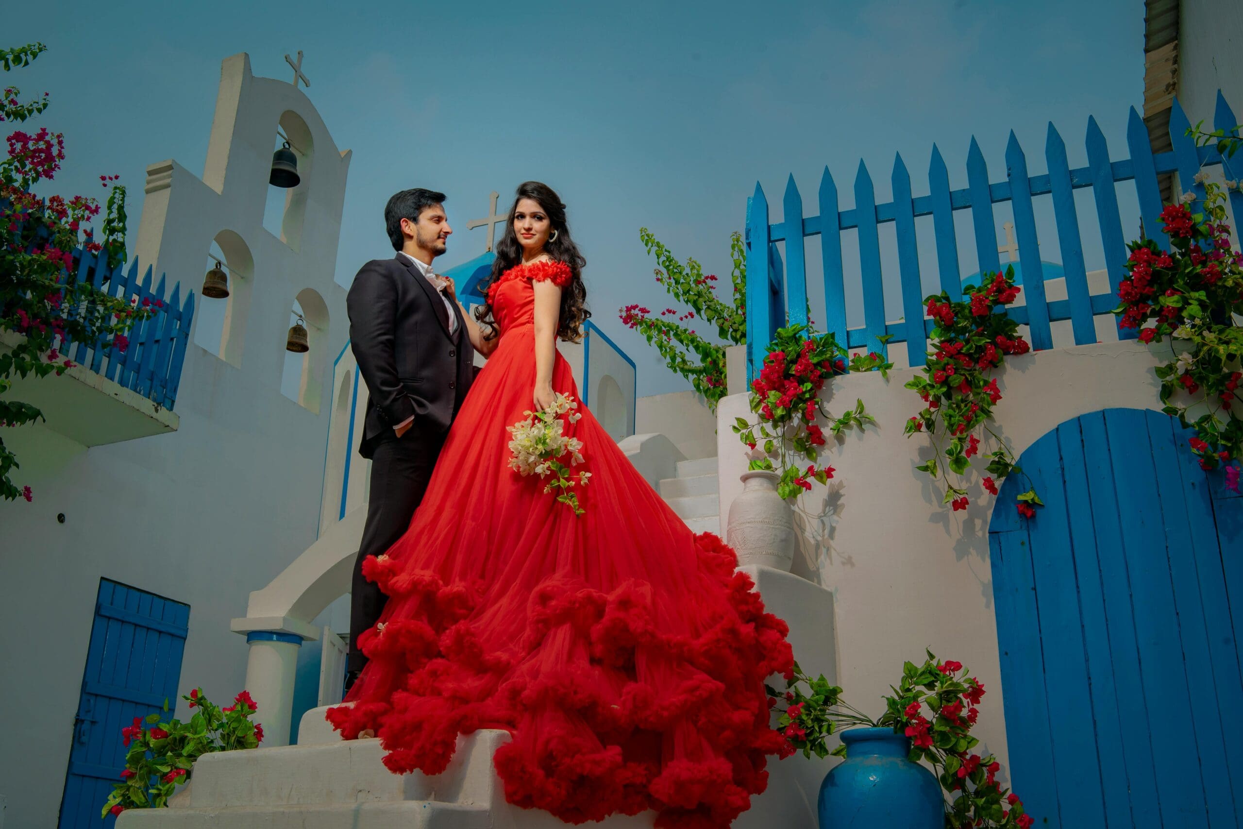 A couple poses for a luxury destination wedding photo in Santorini, Greece. The woman wears a stunning red gown on white steps in front of a whitewashed building with a blue fence, illustrating the "iconic romantic dream" locale.