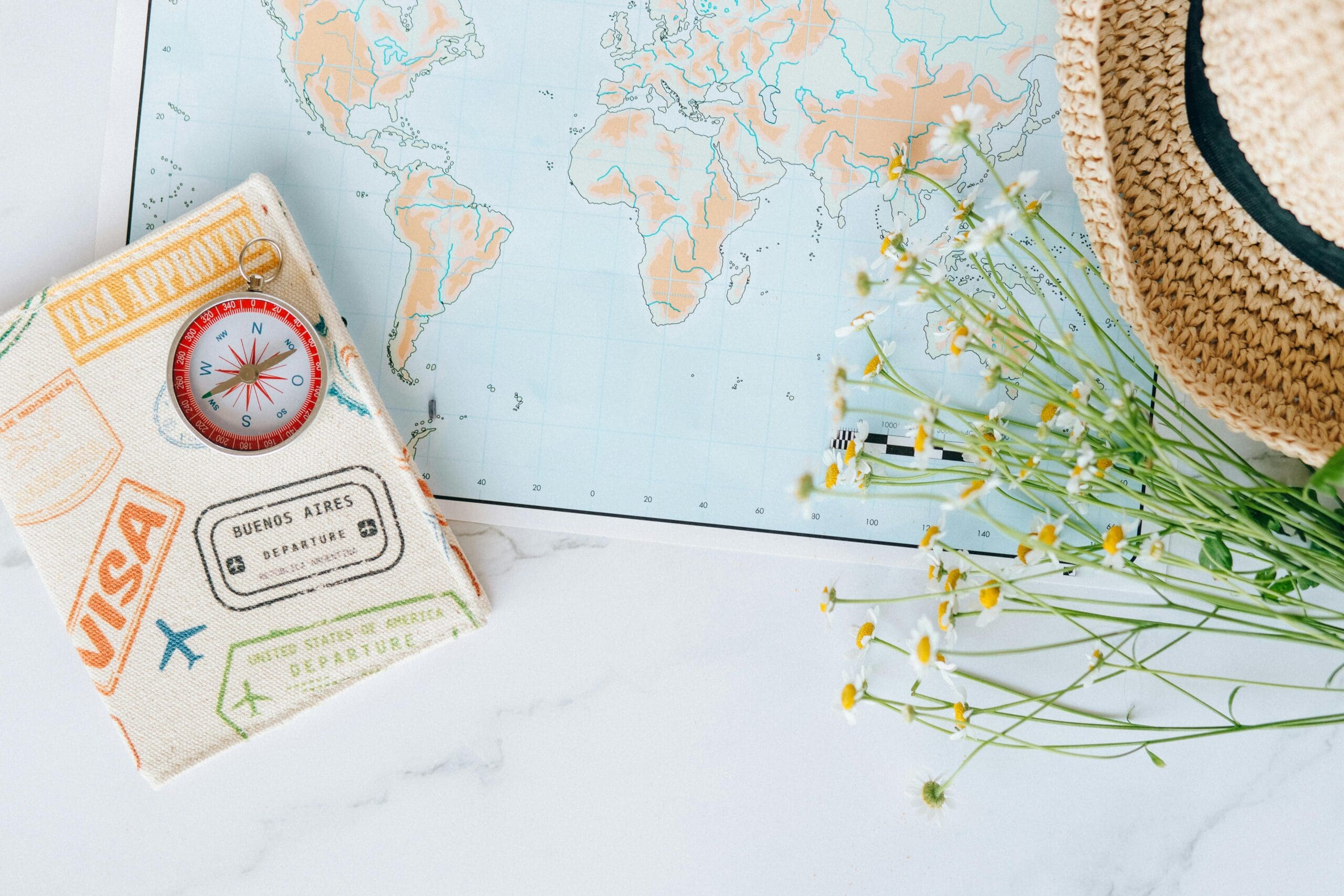 A flat lay of travel items on a white marble surface, including a world map, a passport holder with a compass, a straw hat, and a small bouquet of white and yellow flowers.