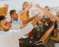 A group of smiling wedding guests in casual clothes clinking their glasses for a toast at a poolside bar during a destination wedding weekend.