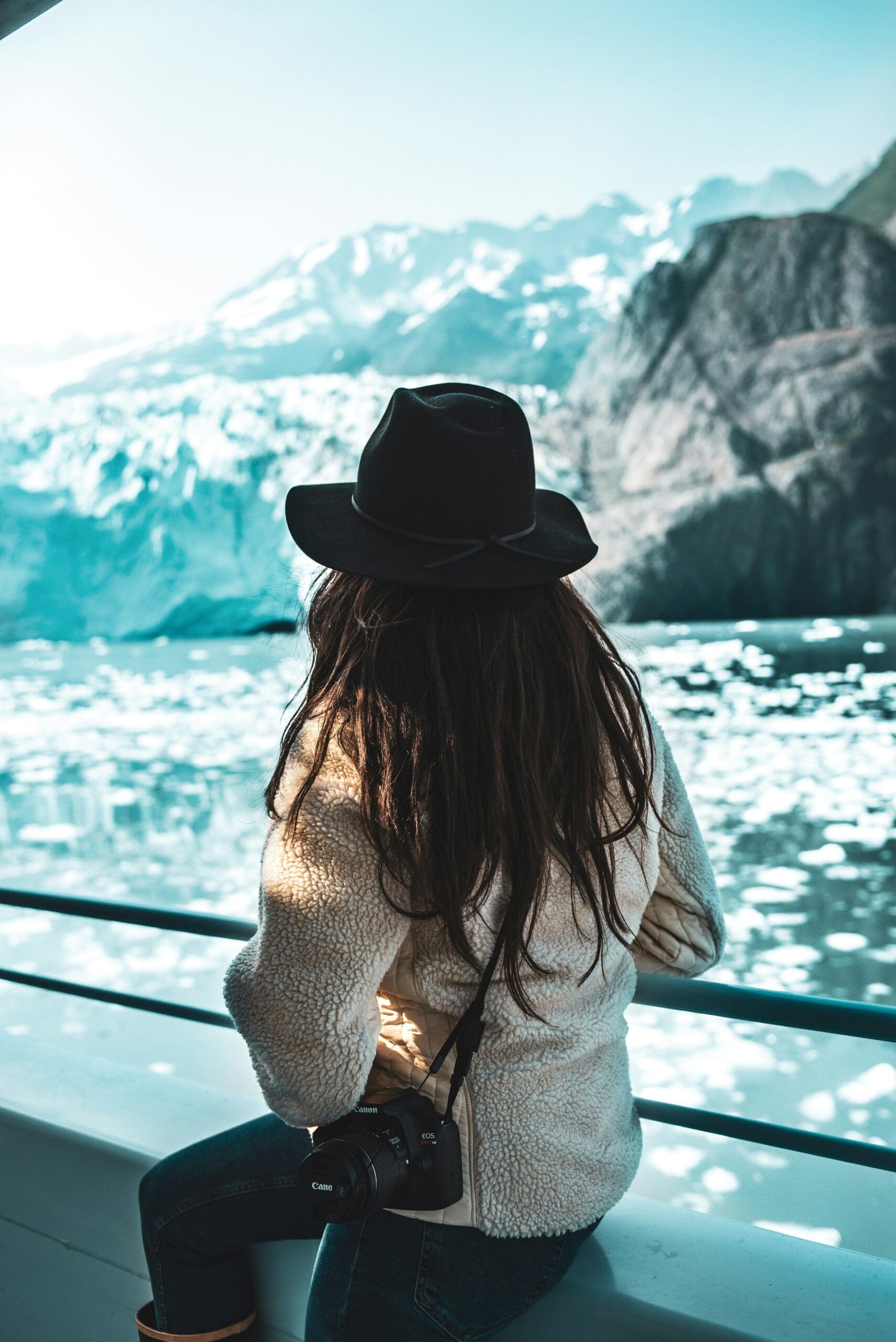 A woman in a black hat and fleece jacket sits on the deck of an expedition boat, looking out at a massive glacier and icy blue water in Alaska.