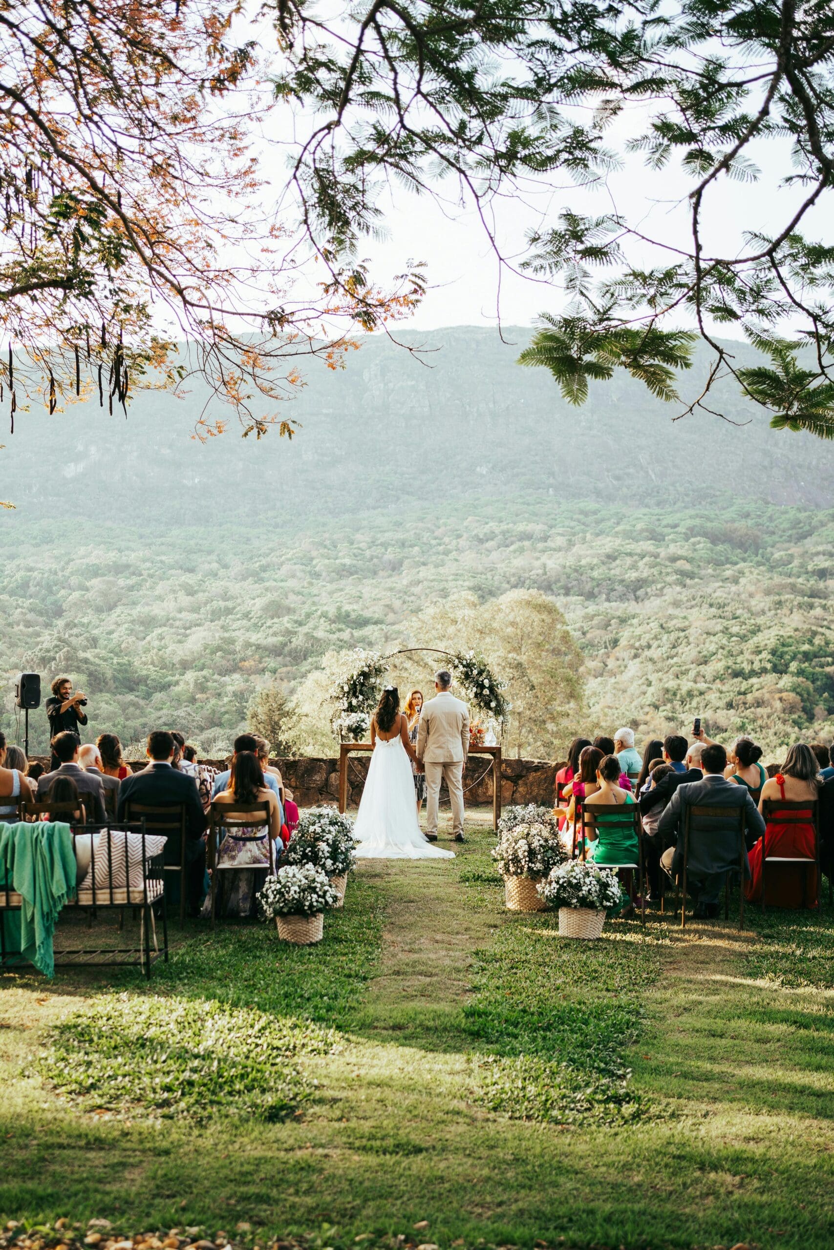 A bride and groom stand at an outdoor altar overlooking a lush green valley during their destination wedding ceremony, with guests seated in the foreground.