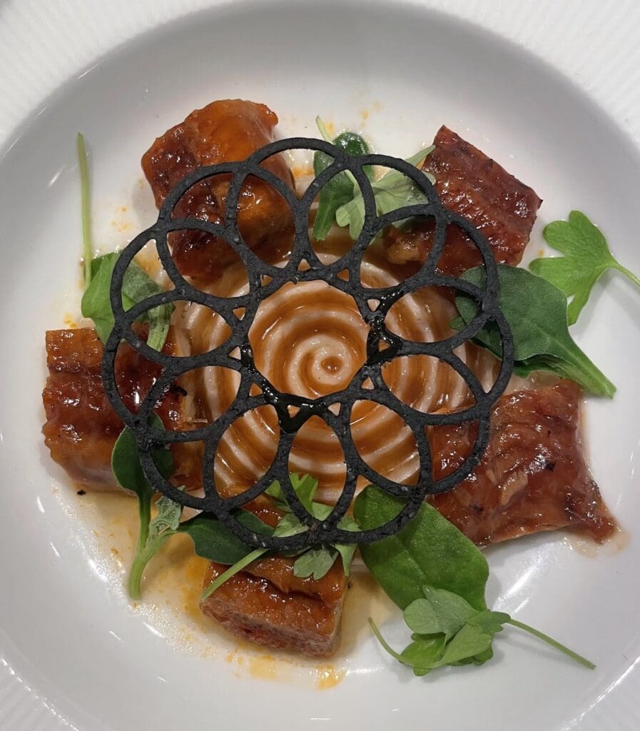 A beautifully plated gourmet dish from a cruise ship's specialty dining restaurant, with a delicate black lattice garnish over a swirl of sauce.